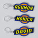 Custom anime-themed DIGIMON keychains with personalized engravings. One keychain reads 'AGUMON' with a blue and yellow font on a black background, and the other reads 'DAVID' with a similar color scheme.